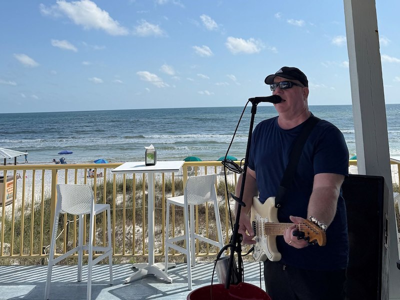 man playing guitar and singing with the beach in the background